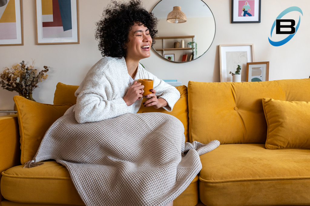 Happy African american woman enjoying quiet time at home laughing, drinking morning coffee sitting on sofa.